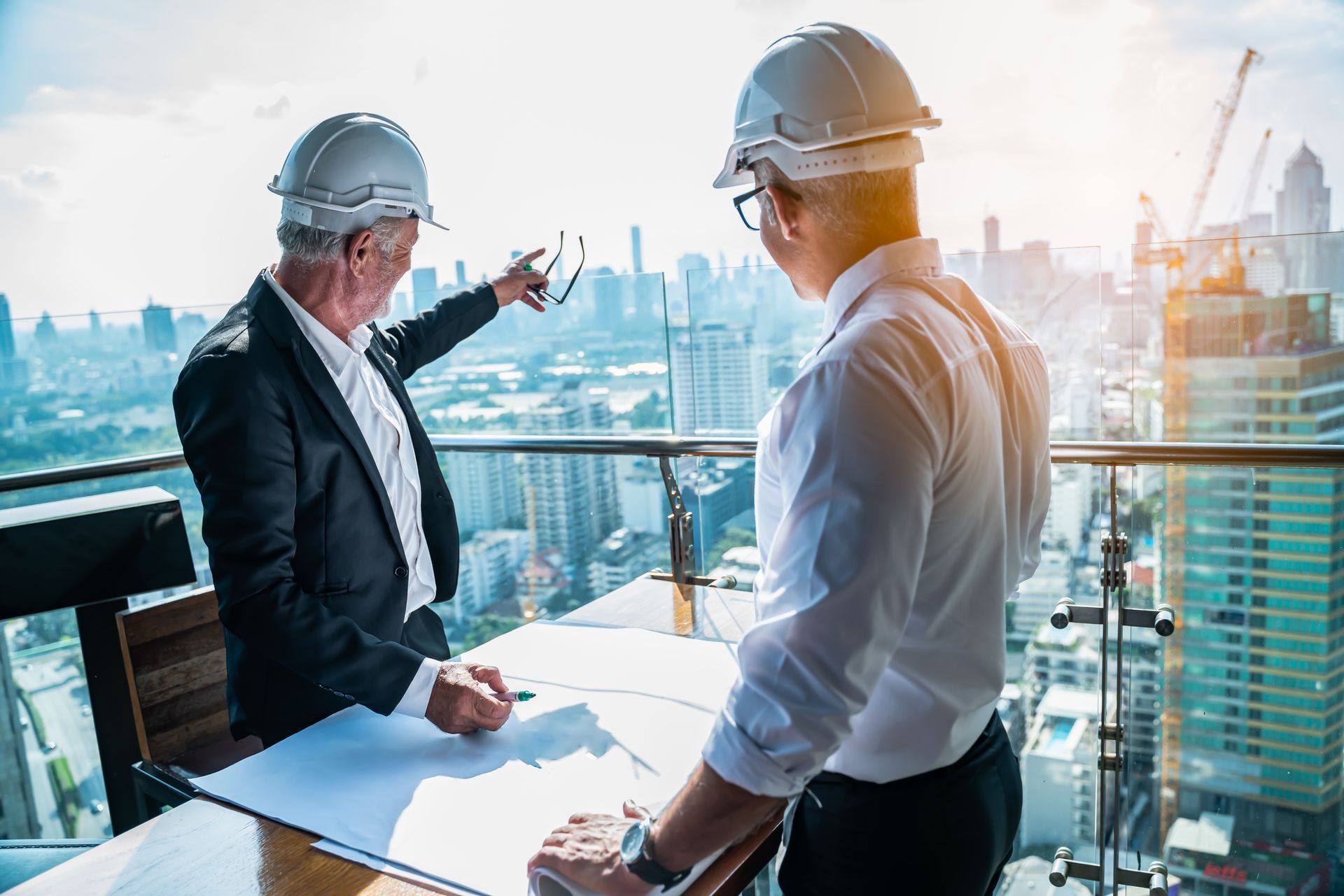 Two engineers in hard hats reviewing blueprints on a balcony overlooking a city skyline