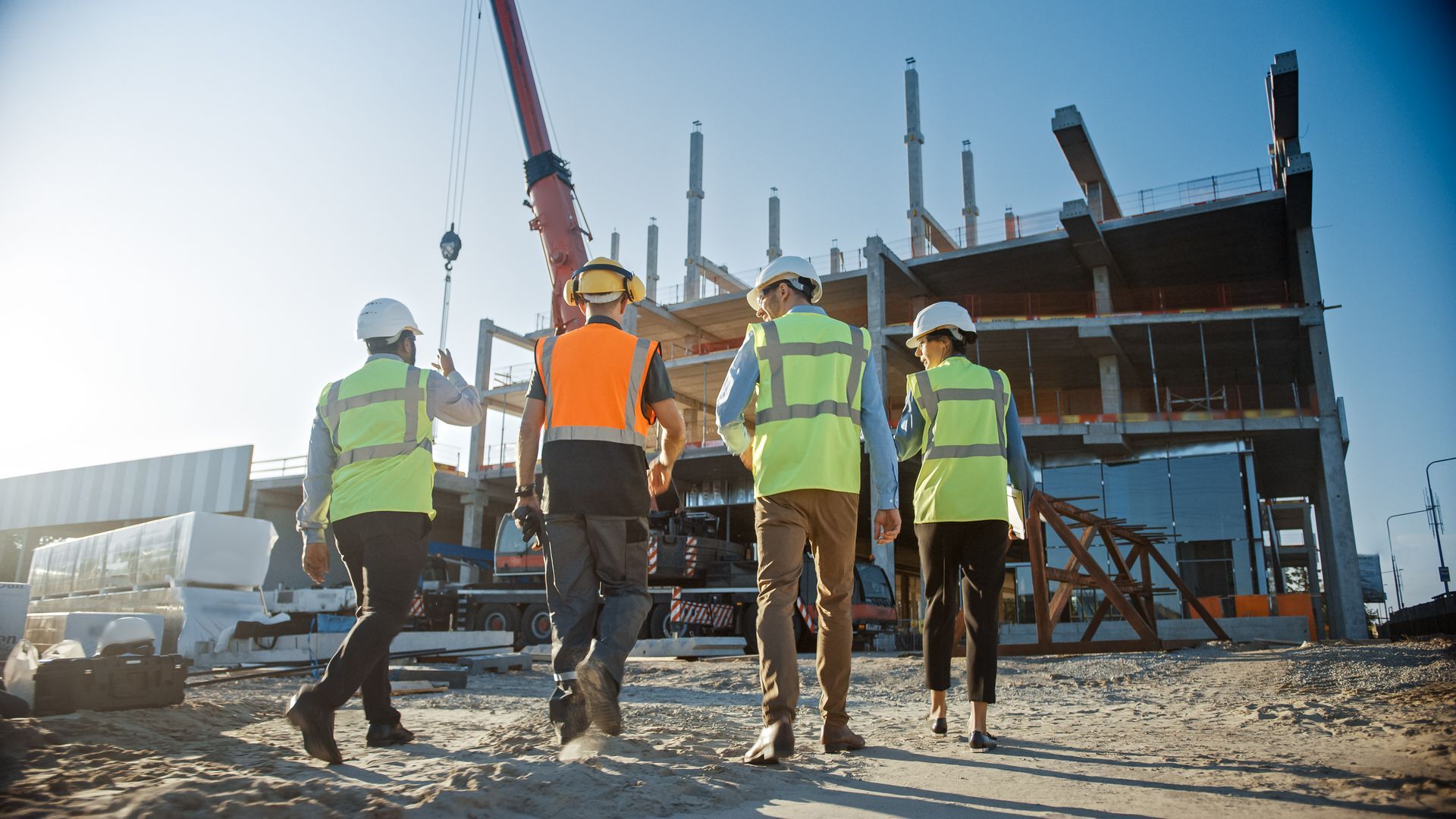 Construction workers in safety vests walking at a building site with a crane and steel frame.