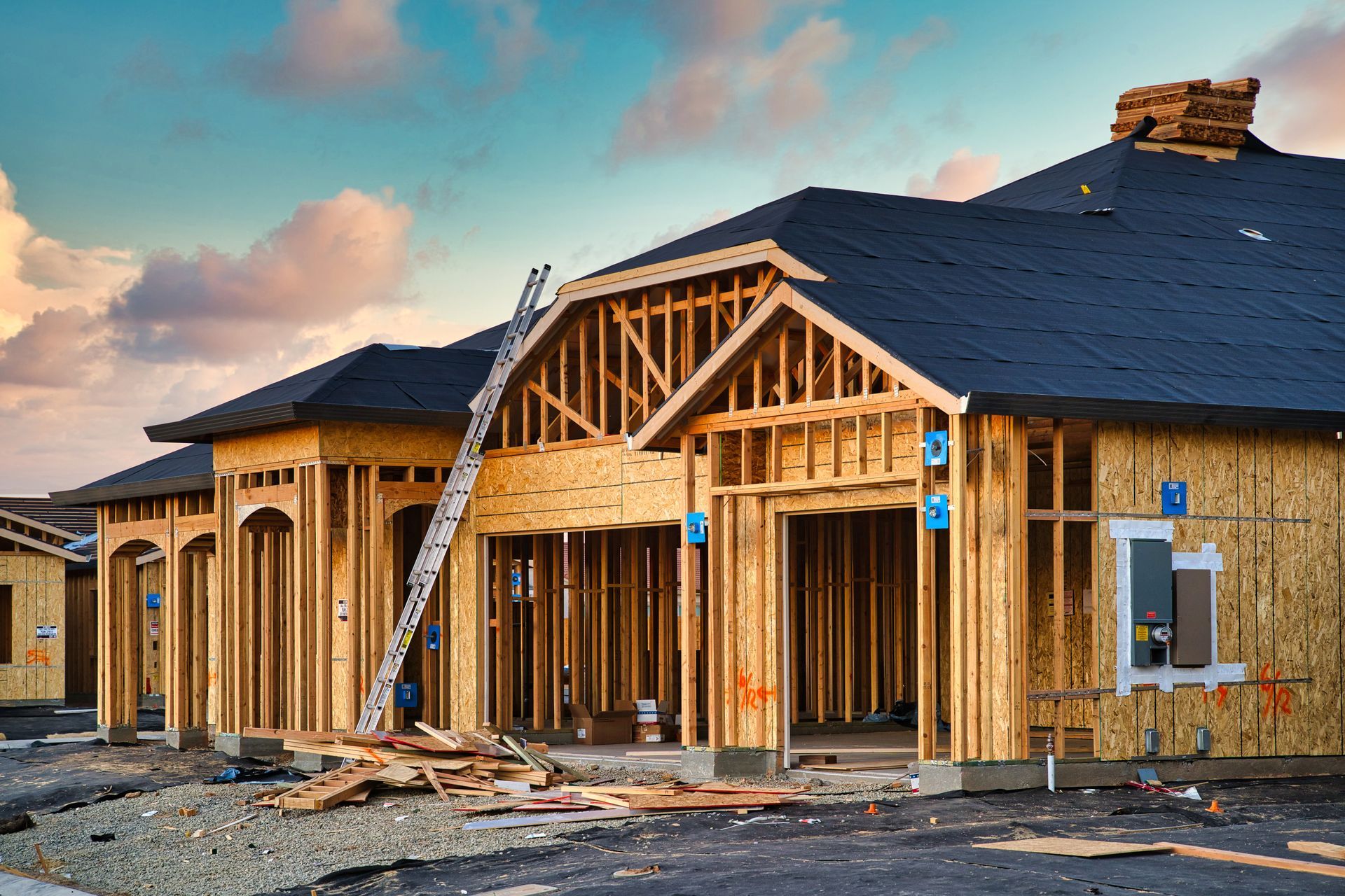 Framed house under construction with exposed wood, ladders, and a partially shingled roof at sunset