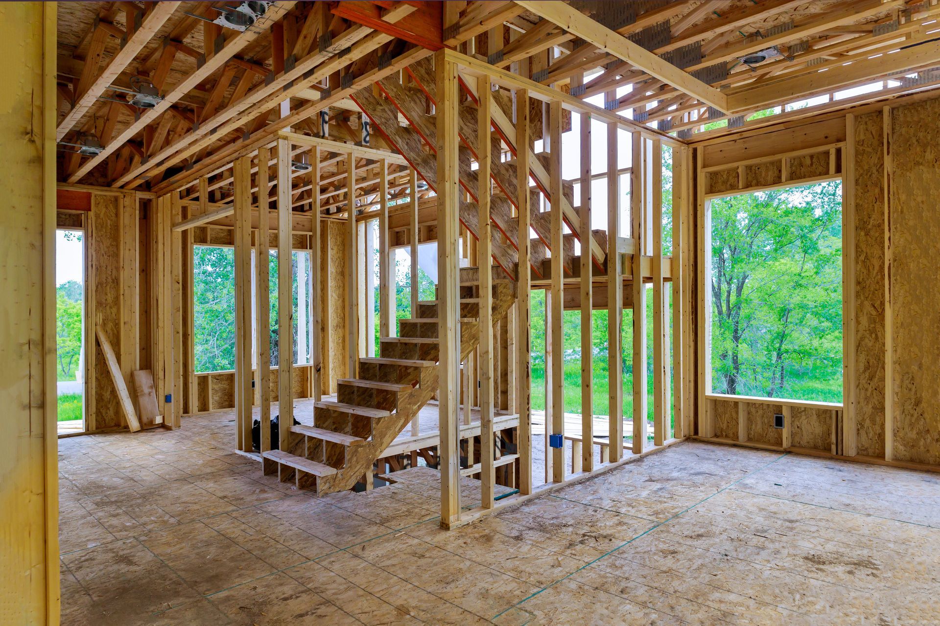 Unfinished house interior with exposed wooden framing, staircase, and large window openings overlooking greenery