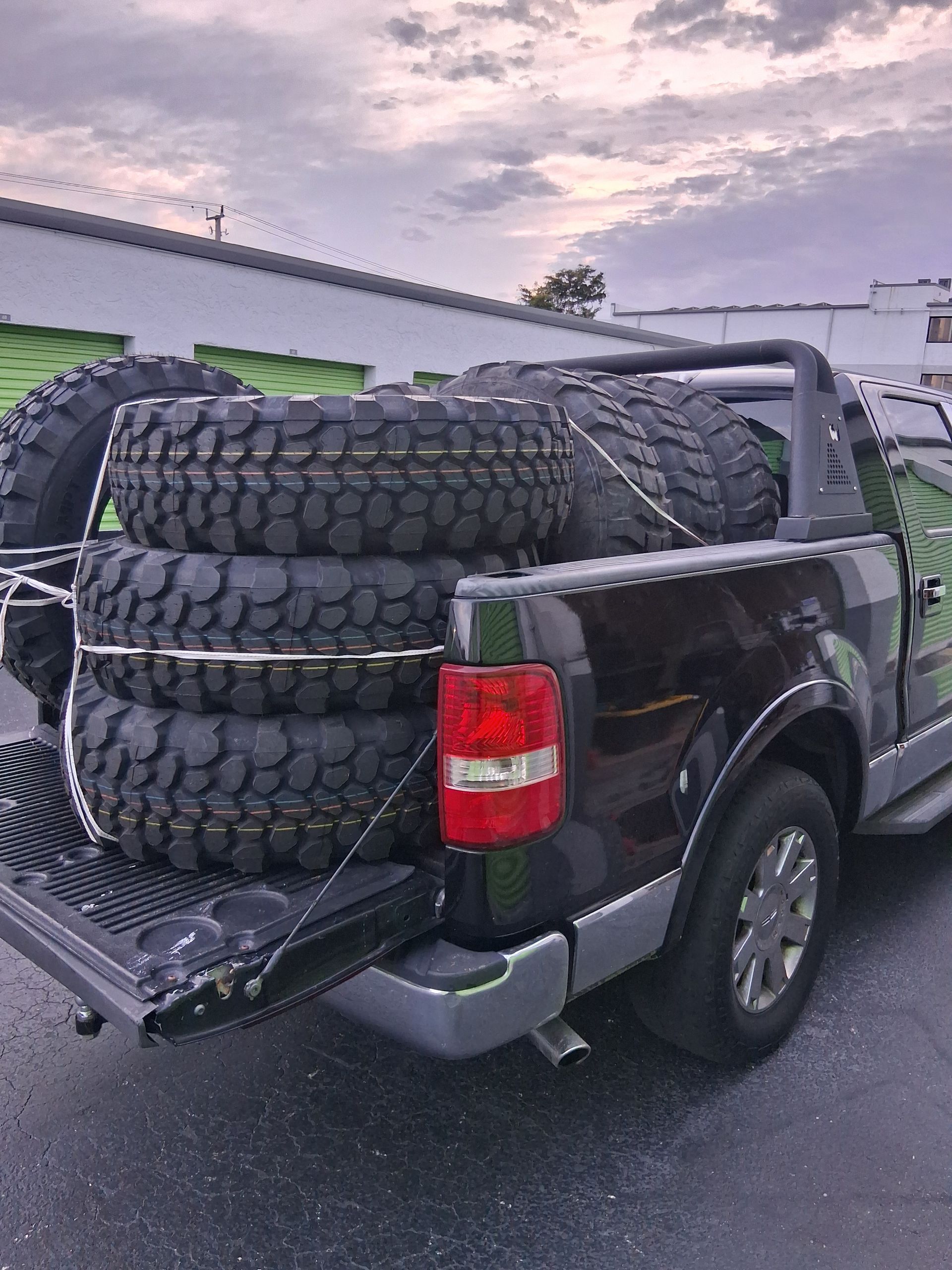 Pickup truck loaded with four large off-road tires in the bed, parked in a lot at dusk.