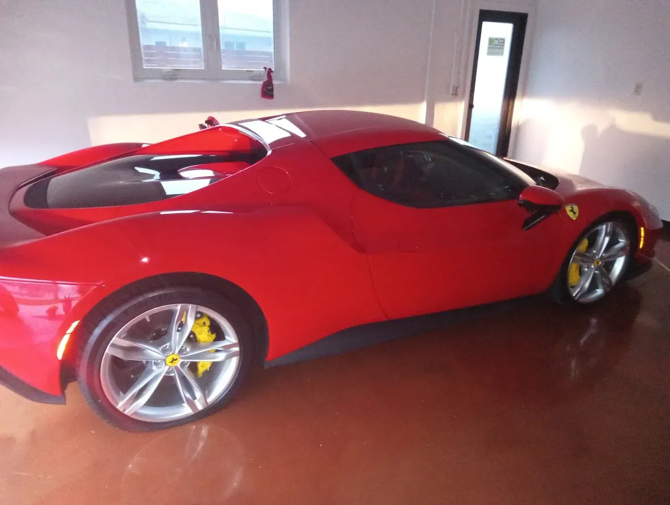 Red sports car parked indoors in a garage with white walls and a window.
