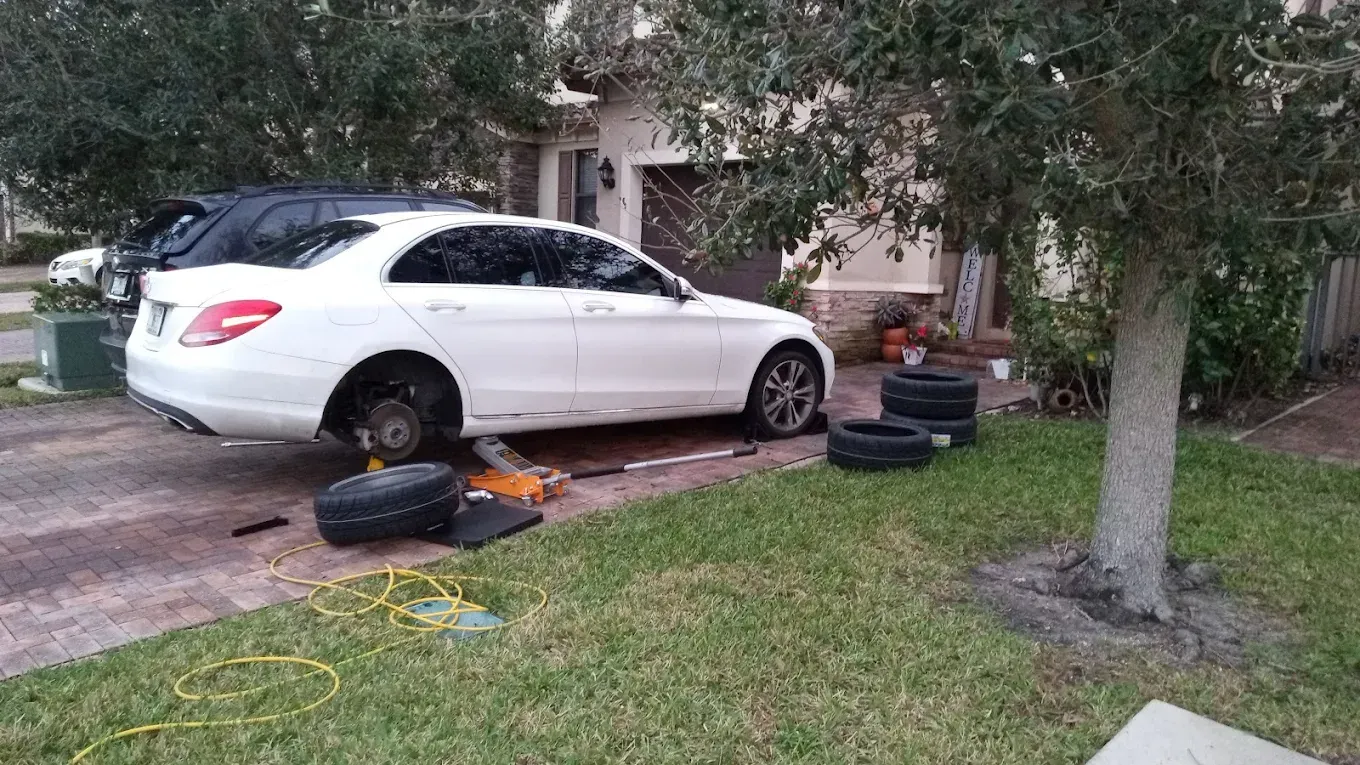 White sedan parked on a driveway with scattered car parts and tools beside it, near a tree and lawn.