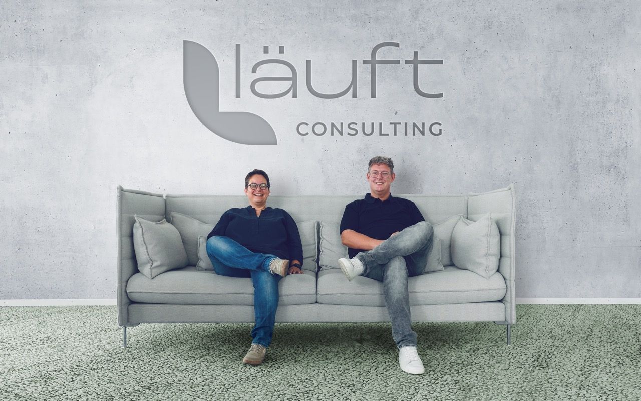 Elke und Jens lächelnd auf einer Couch vor einer Betonwand mit dem Logo von läuft Consulting