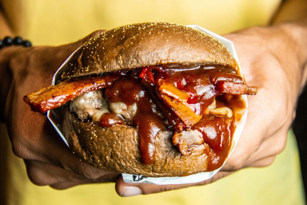 A person is holding a hamburger with bacon and ketchup in their hands.