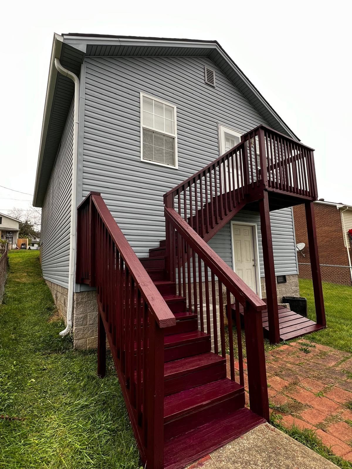 Rear view of a gray house with maroon stairs and an elevated deck, beside a grassy yard and brick path