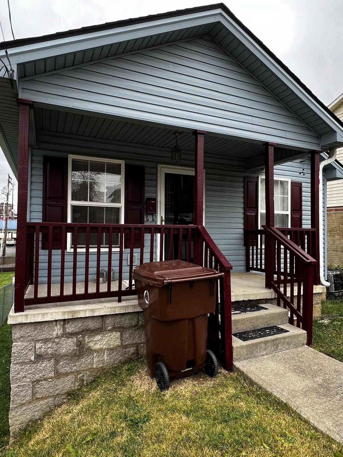 Small gray house with red porch railing and steps, a brown trash bin in front, and a narrow walkway.