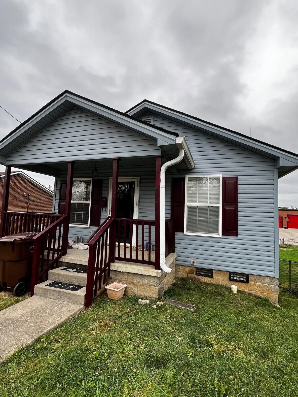 Small blue house with burgundy trim, front porch, and steps under a cloudy sky.