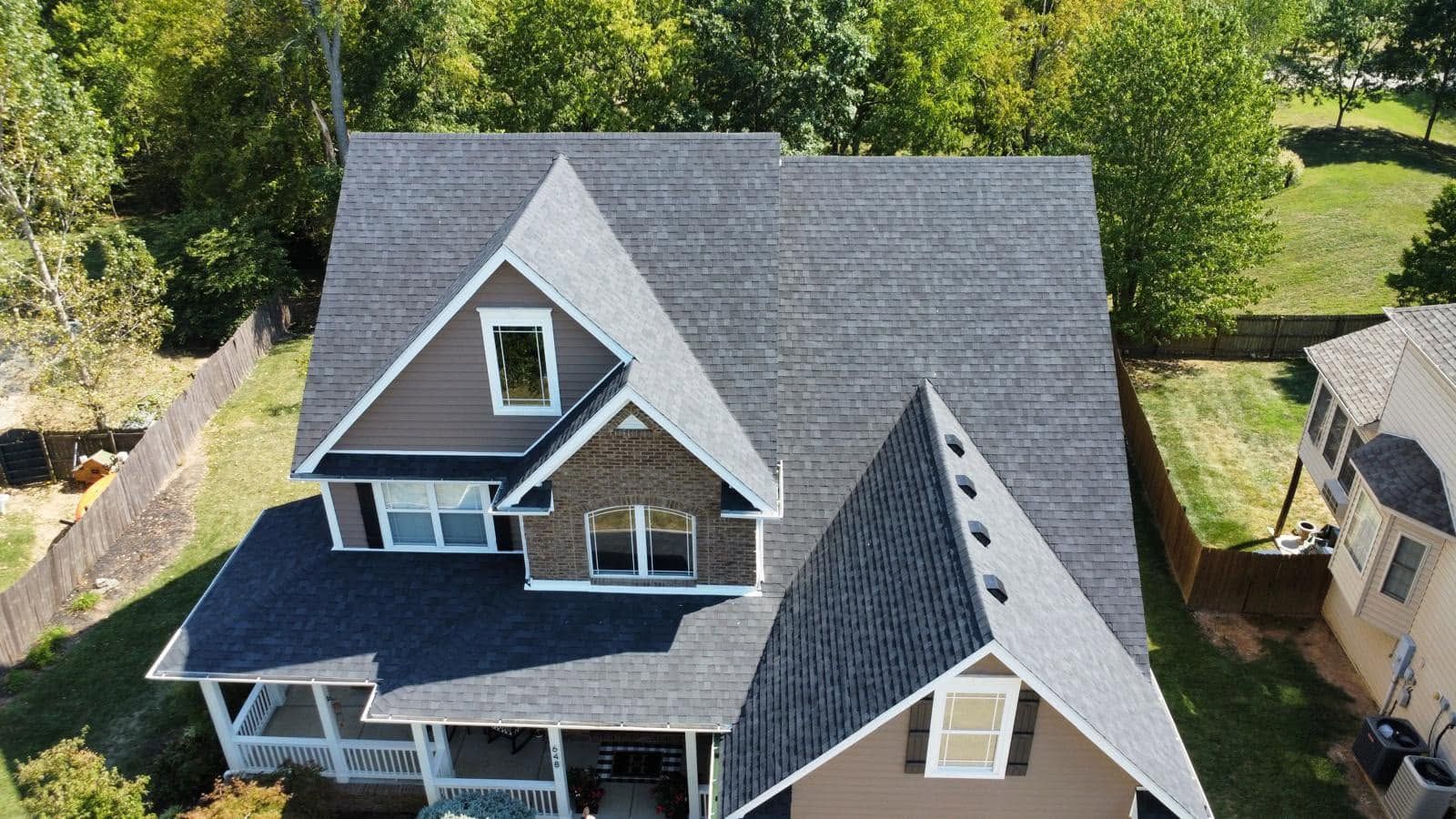 Aerial view of a gray-roofed house with dormer windows and a front porch, surrounded by trees and greenery