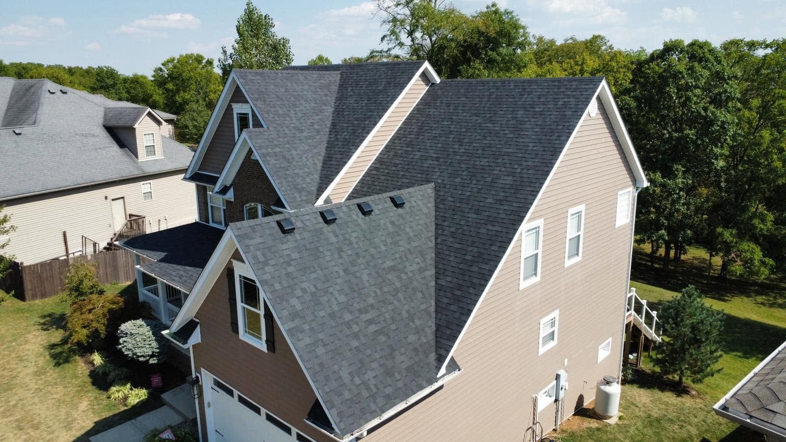 Aerial view of a tan house with a dark gray roof and dormers, surrounded by trees and neighboring homes