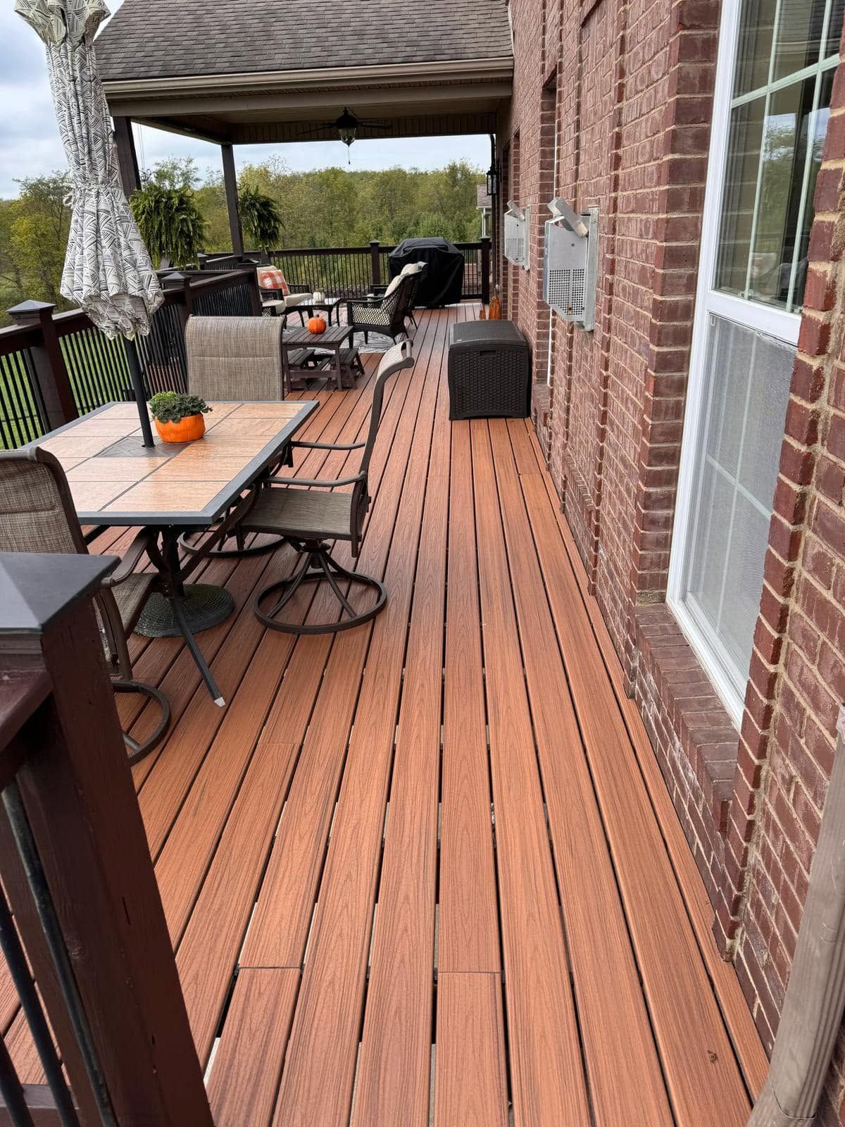 Brick patio deck with outdoor dining table, grill, chairs, and potted plants under a covered awning.