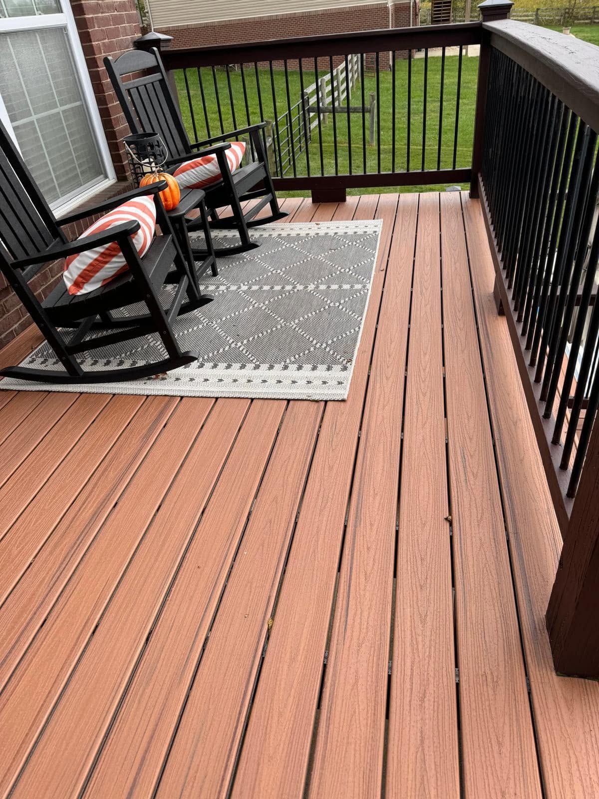 Wooden deck with two black chairs on a gray rug, overlooking a grassy yard with a black railing.