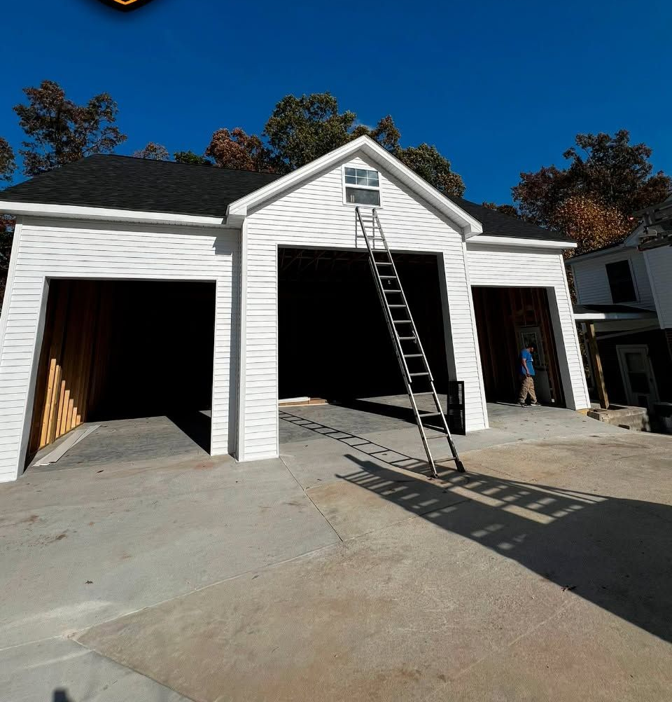 White three-bay garage under construction with open doorways and a ladder leaning to the center gable.