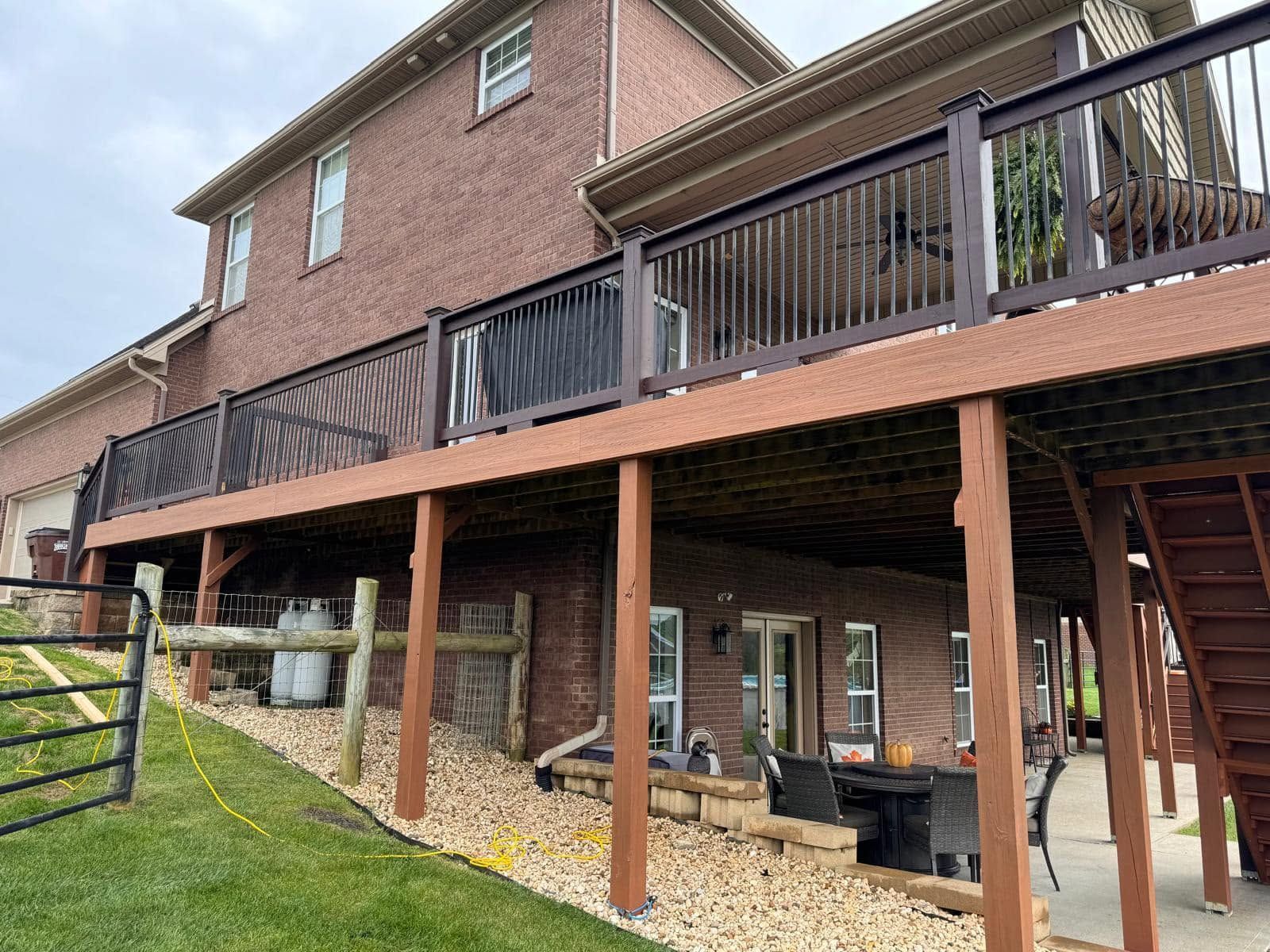 Brick house with raised wooden deck, stairs, and a gravel-covered patio beneath.