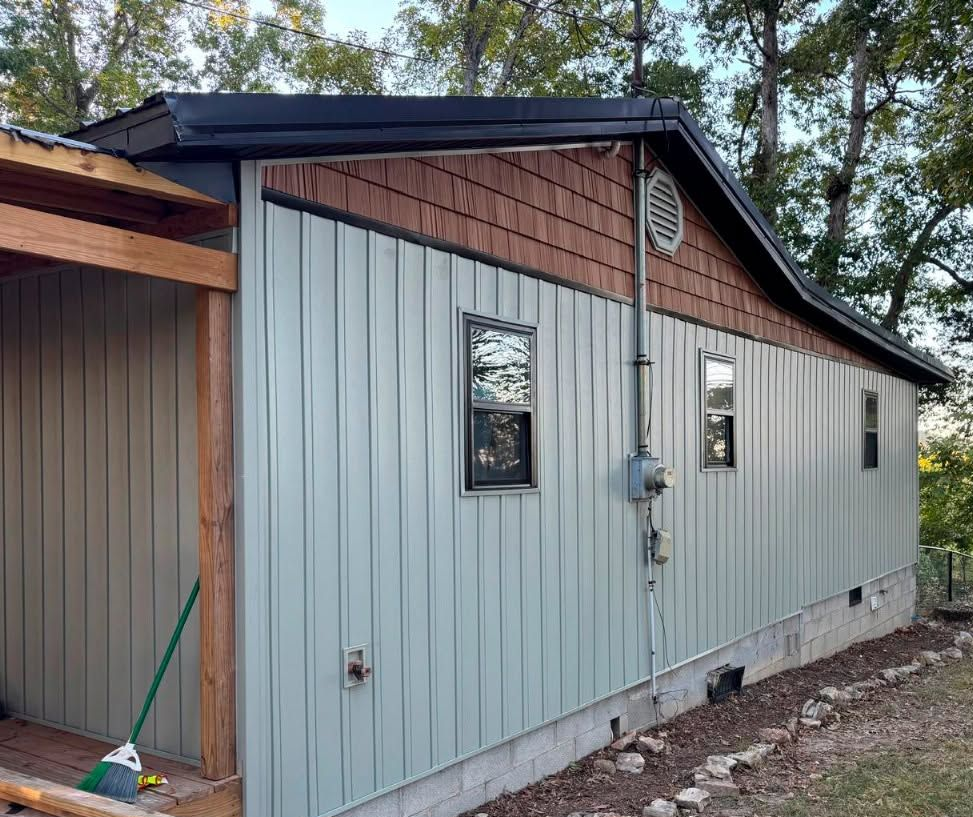 Gray metal-sided building with a black roof and small windows beside a wooden structure outdoors