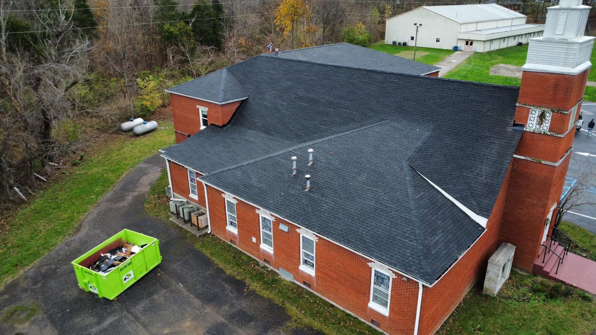 Aerial view of a red brick church with a black roof and tall brick tower, beside a green dumpster and parking lot