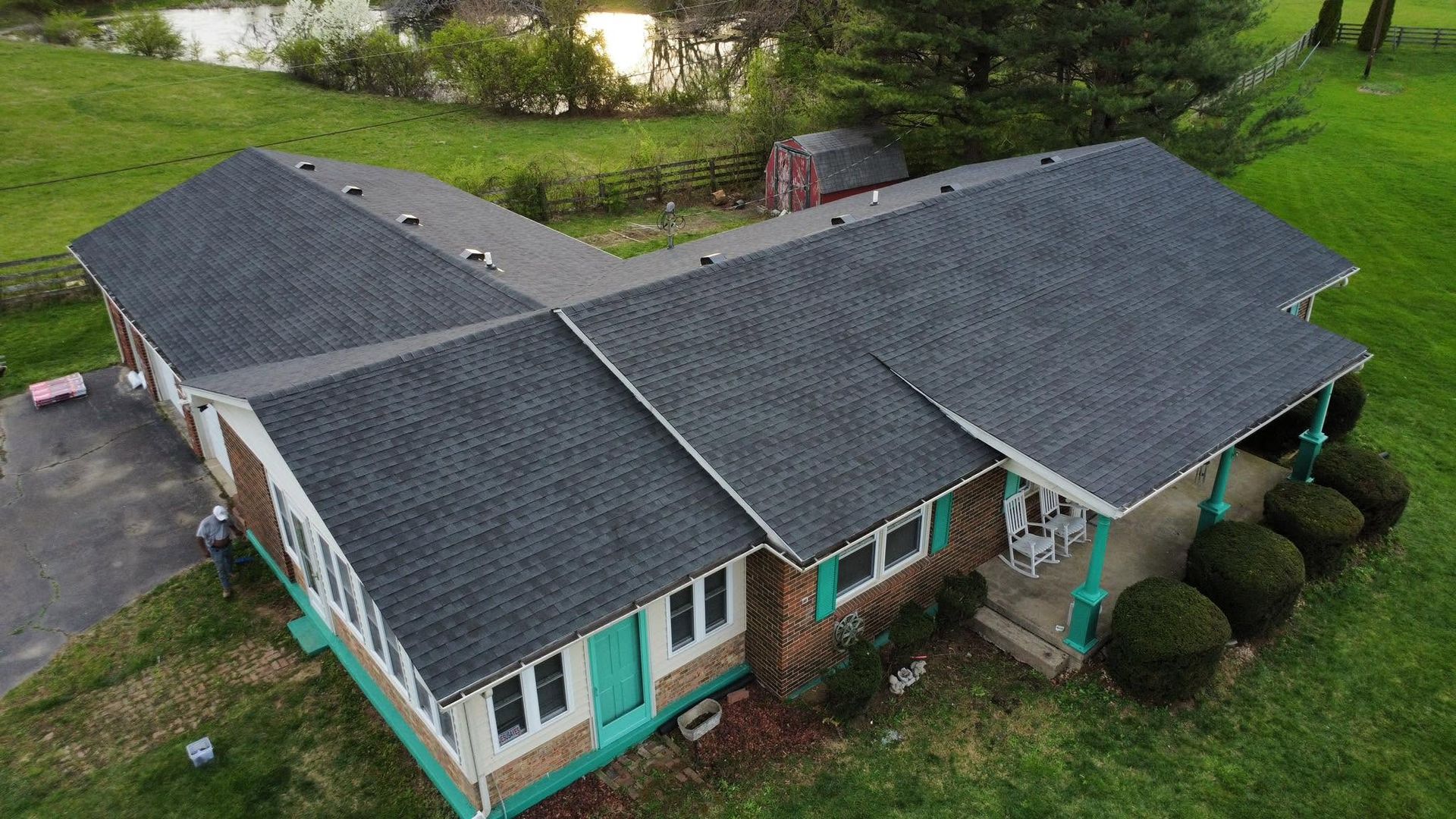 Aerial view of a single-story house with a dark gray roof and teal trim surrounded by green lawn and trees