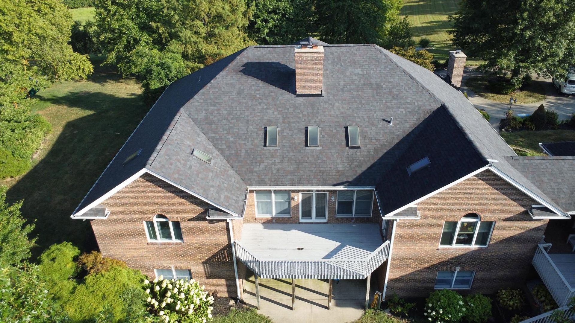Aerial view of a large brick house with a gray roof and central balcony, surrounded by trees.
