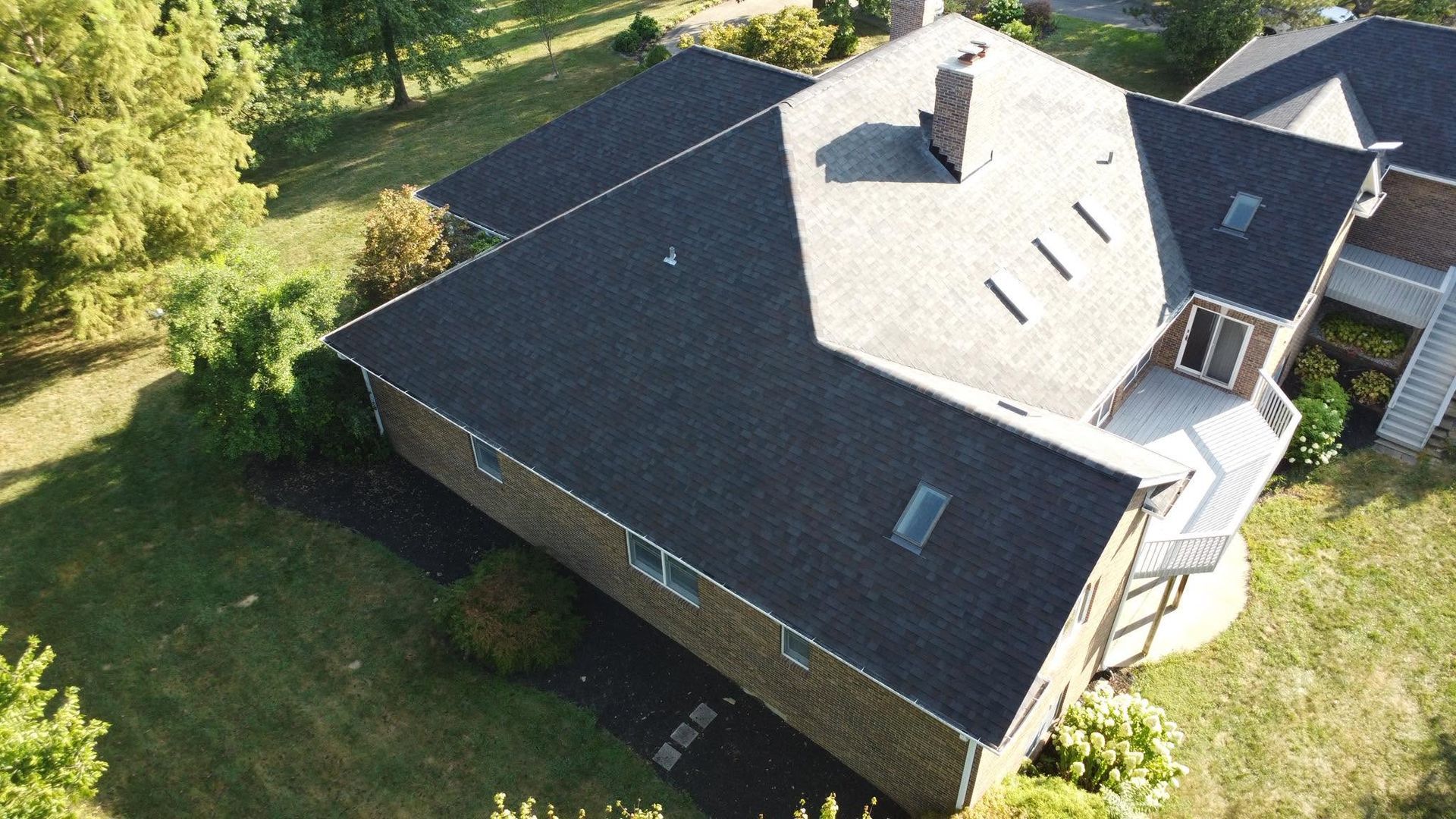 Aerial view of a house with a dark shingled roof, skylights, and a small deck, surrounded by green lawn and trees