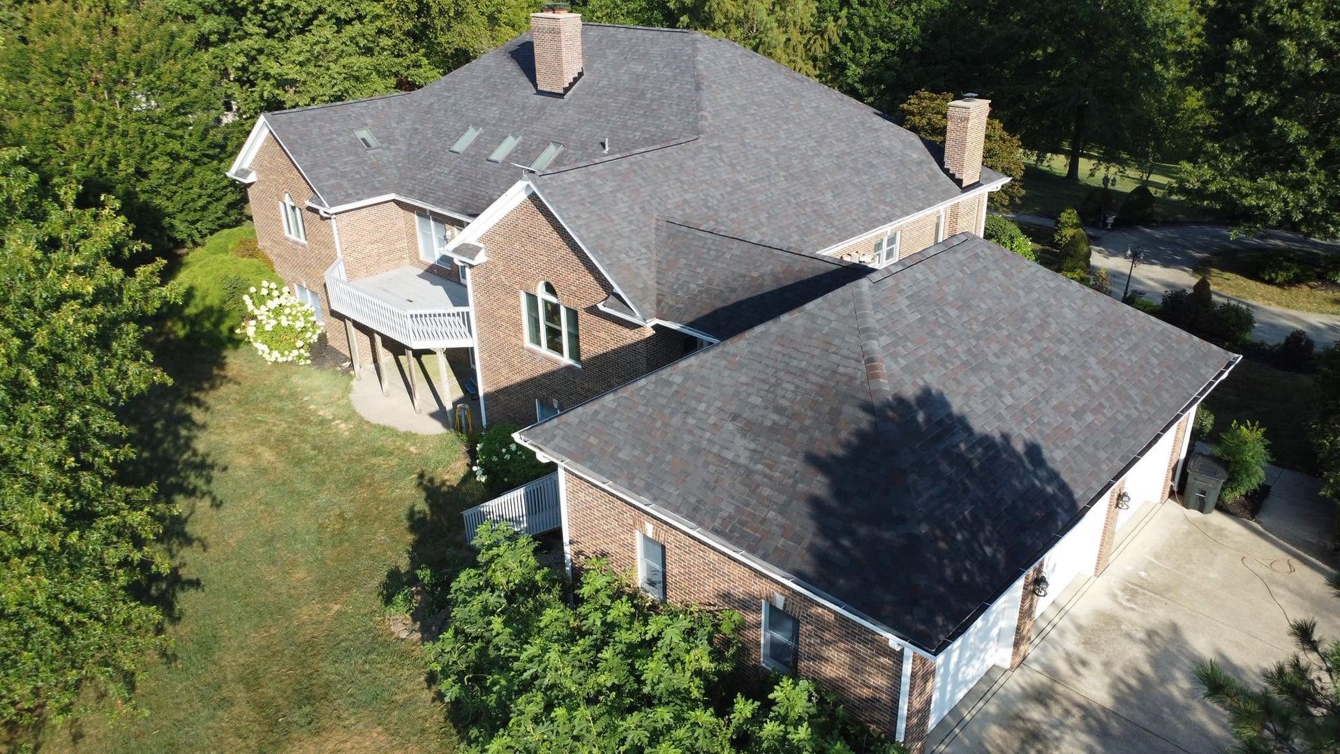 Aerial view of a brick suburban house with gray roof, driveway, and trees surrounding the property