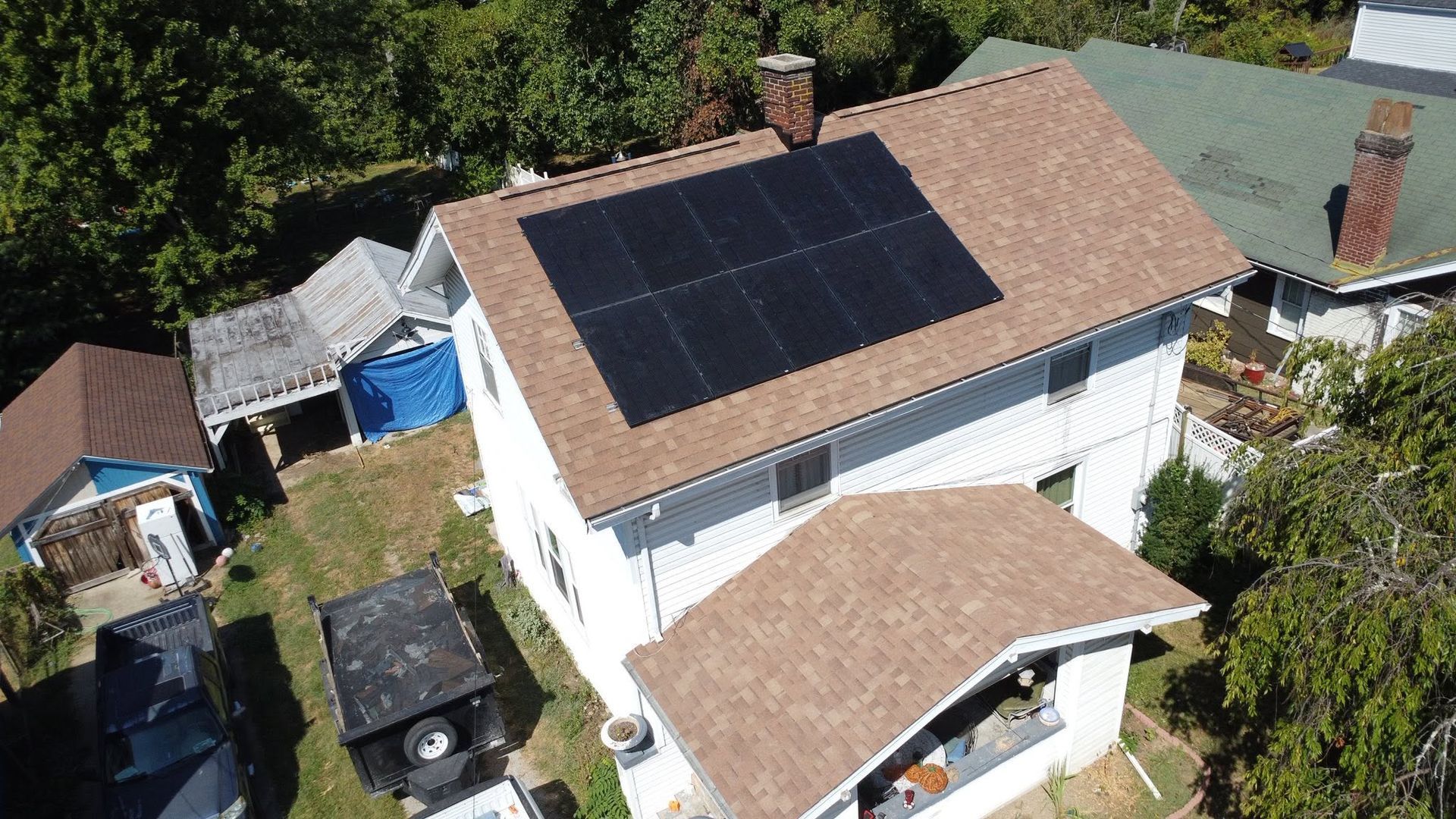 Aerial view of a white house with a tan roof and large solar panels, surrounded by trees and a driveway