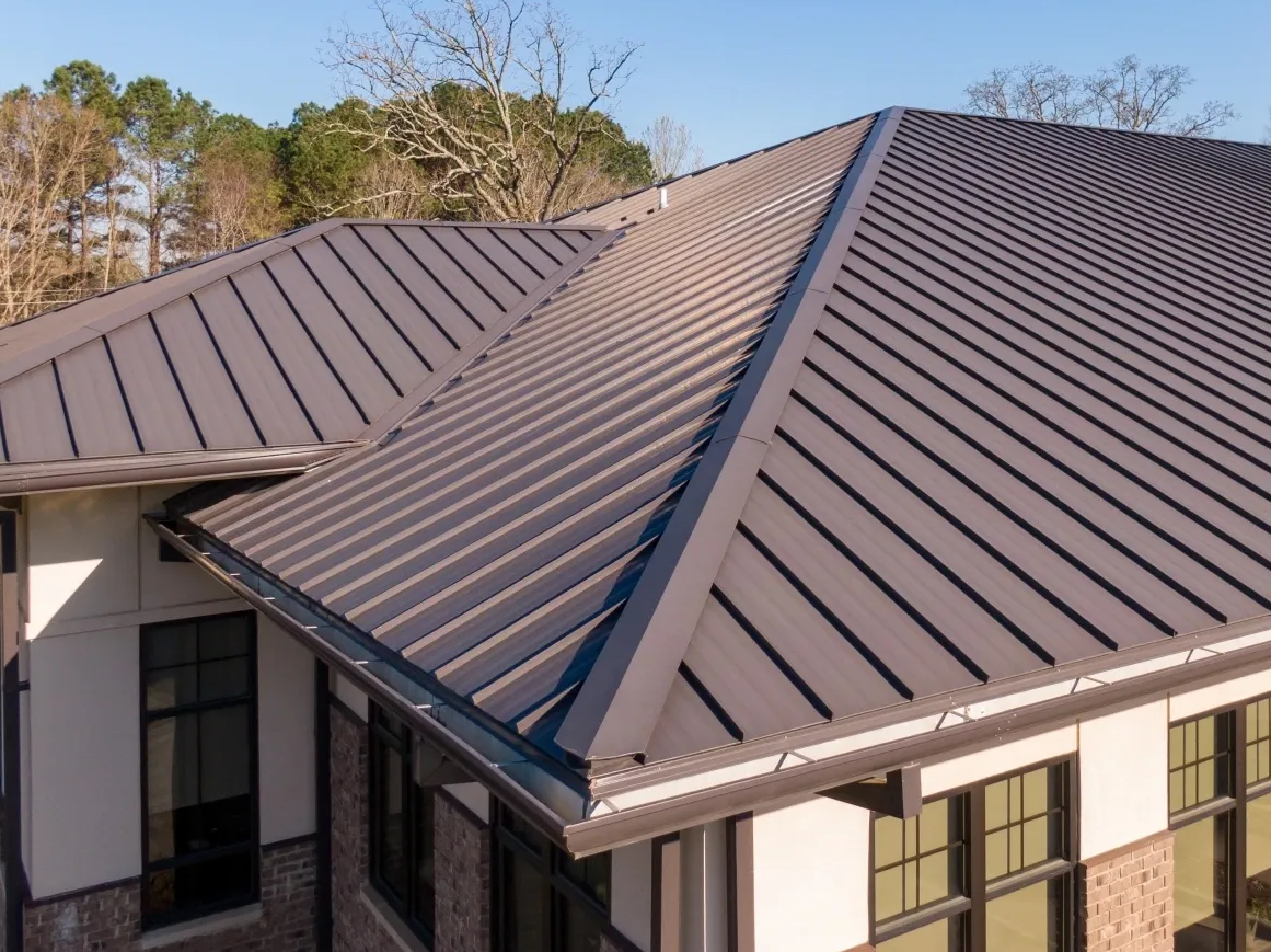 Corner of a house with a dark gray metal roof and beige siding, surrounded by trees