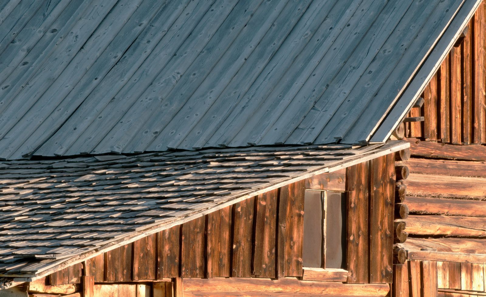 Weathered wooden barn with a gray metal roof, showing a close-up of the exterior wall and roofline.
