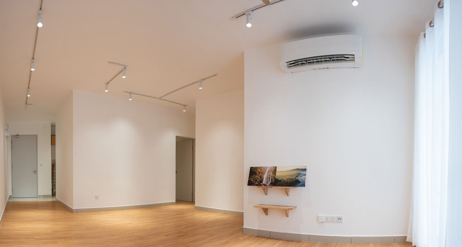 Spacious modern room with white walls, wood floor, ceiling lights, and large windows with sheer curtains.