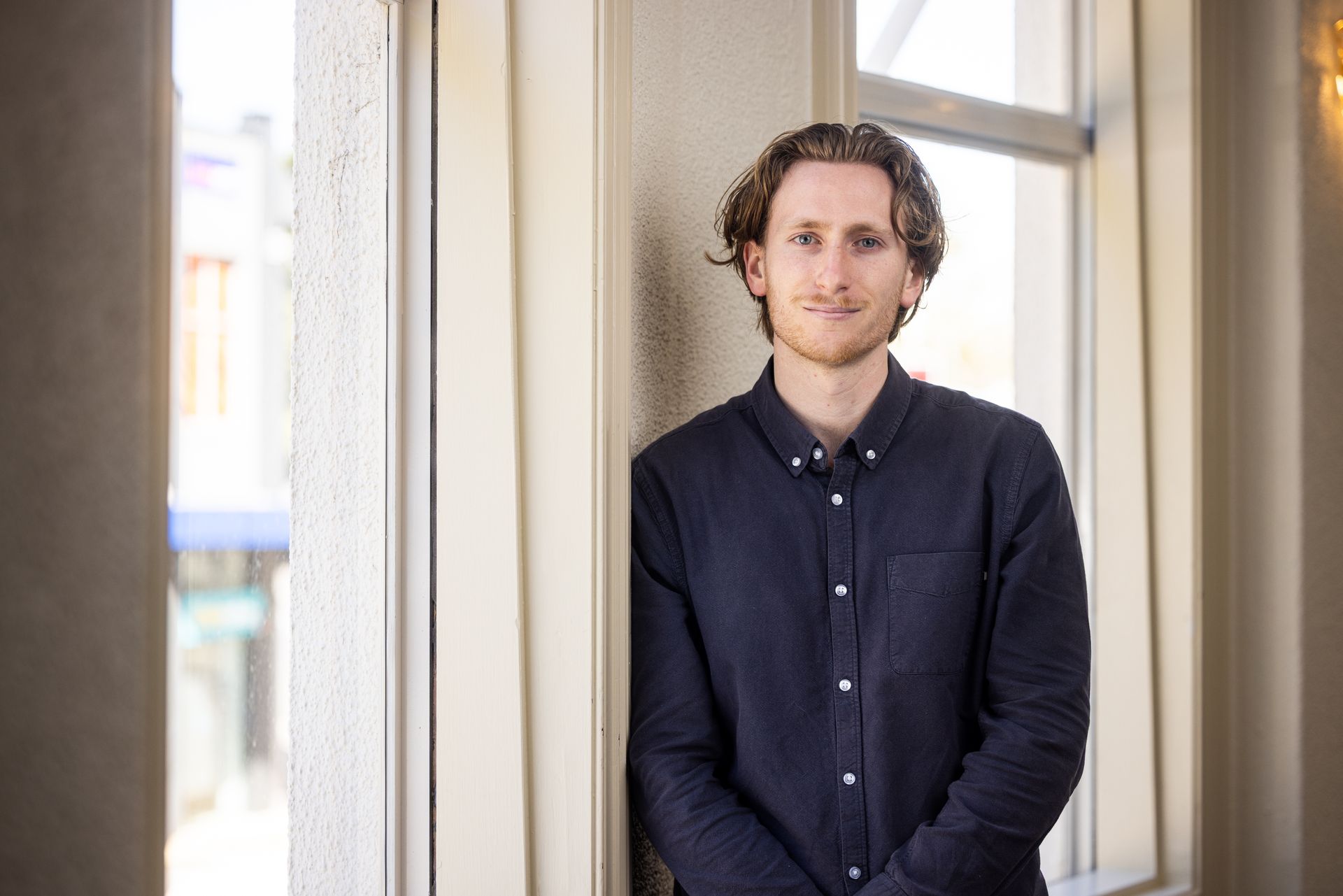 Person standing in a bright hallway between windows, wearing a dark button-up shirt.