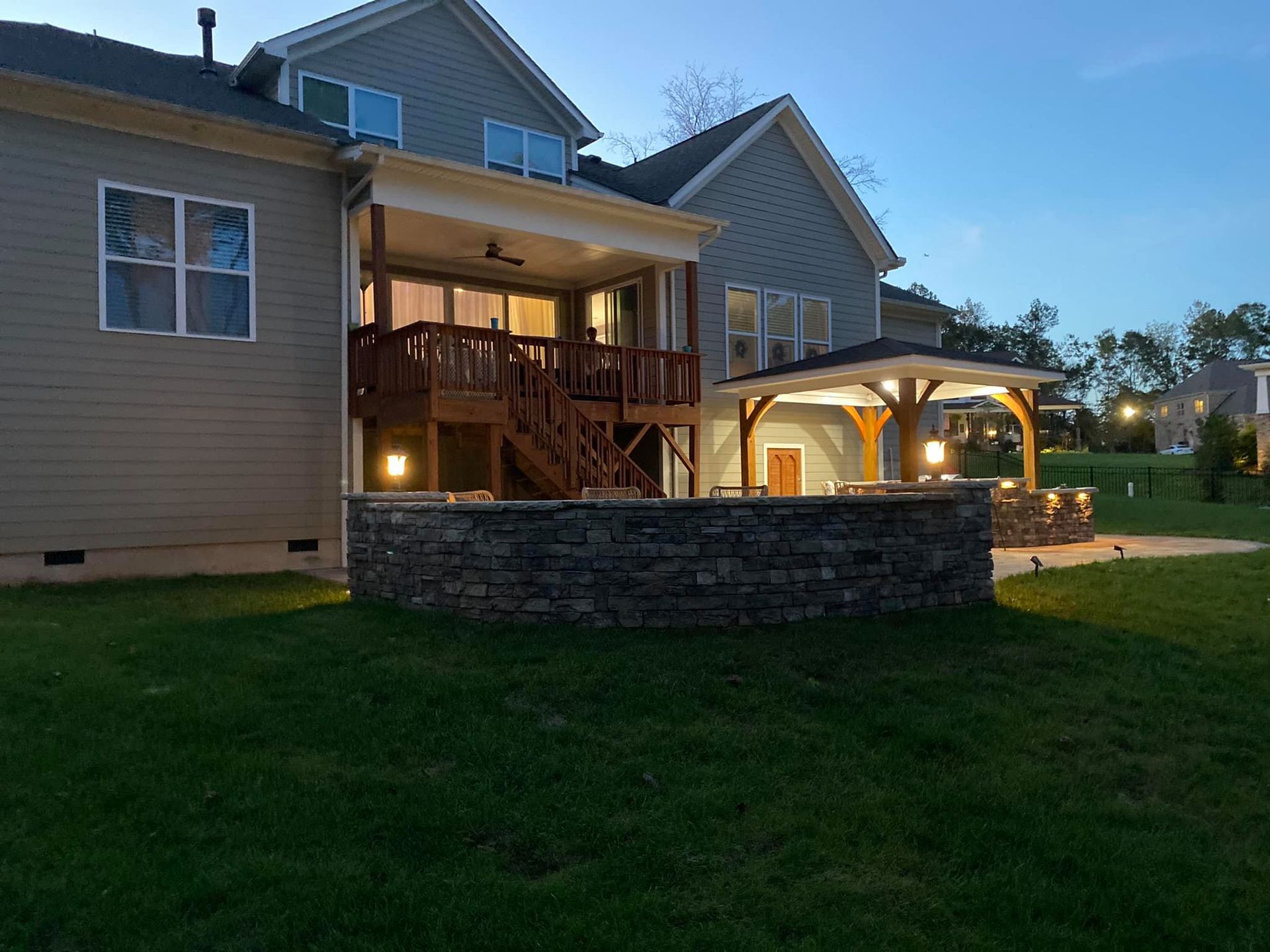 Backyard with a stone wall patio, deck, gazebo, and house at dusk.