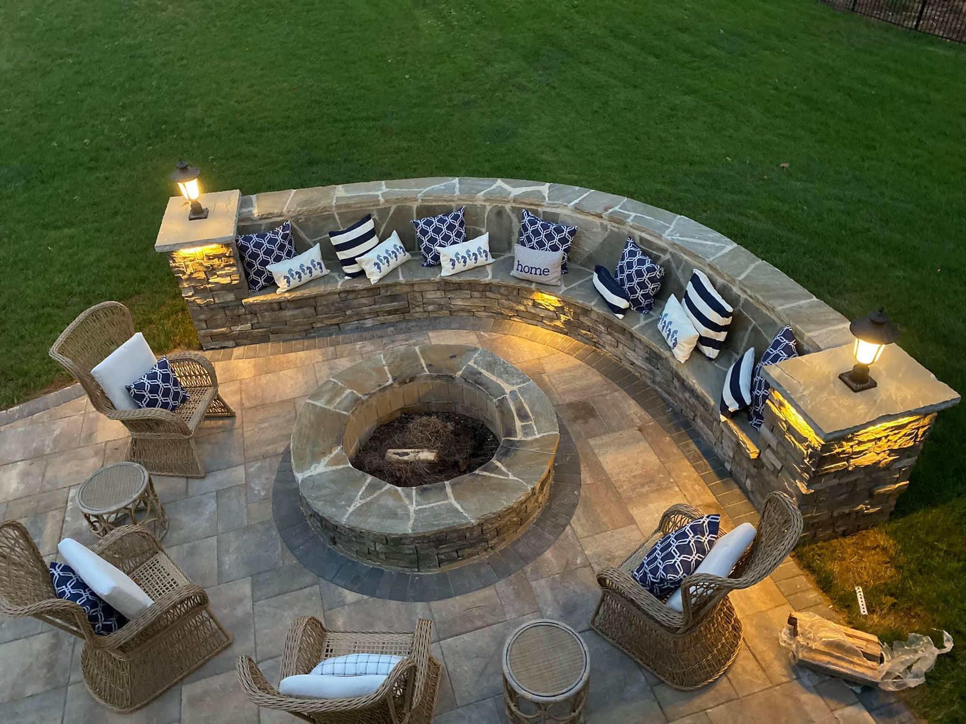 Stone fire pit surrounded by curved stone seating with blue and white pillows. Wicker chairs on a patio with grass.