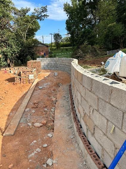 Curved cinder block wall under construction in a yard with red soil and brick piles.