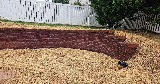 Brick retaining wall with steps, curving along a dry, grassy yard, white picket fence in background.