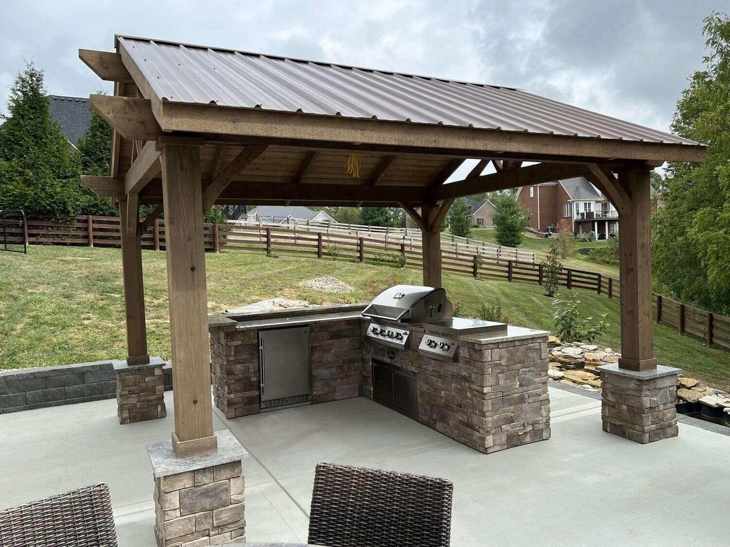 Outdoor kitchen with stone counters, grill, and metal-roofed wooden pergola on a concrete patio.
