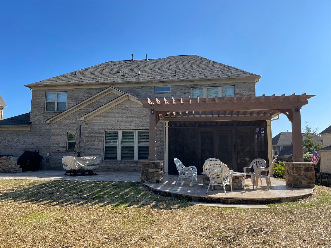Backyard with brick house, pergola over patio with seating. Sunny day, dry grass.