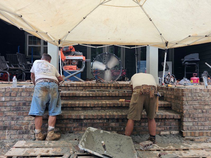 Two workers constructing brick steps under a tent. A large fan blows. Construction materials are visible.