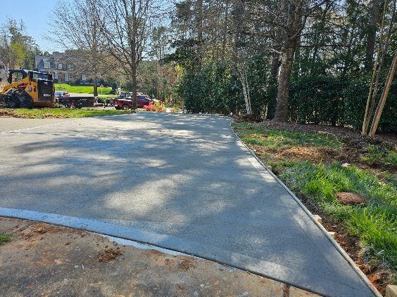 Newly poured concrete driveway with a curb, adjacent to grass and trees, construction equipment in the background.