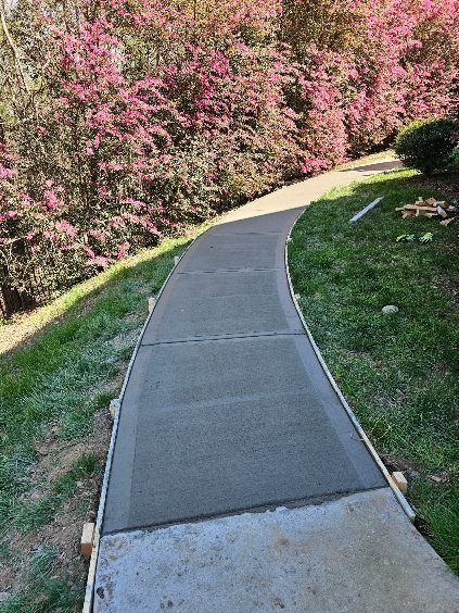 Newly poured concrete sidewalk curves uphill, bordered by grass and flowering bushes.