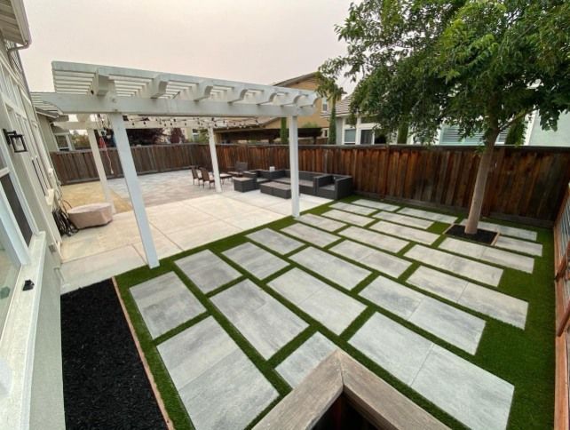 Backyard patio with pergola, seating area, large pavers with artificial grass, and a wooden fence.
