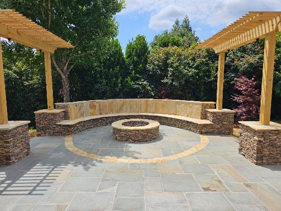Stone patio with fire pit, built-in curved seating, and two wooden pergolas; lush green trees in background.