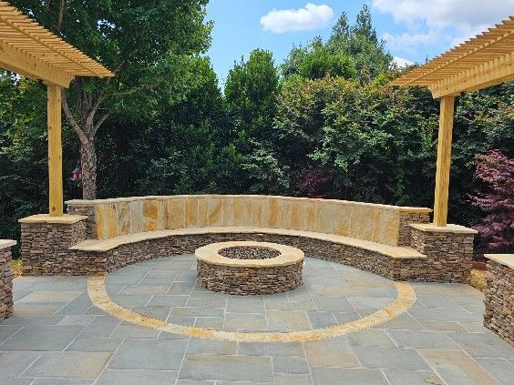 Stone fire pit and curved bench in a backyard patio with pergolas and lush greenery.