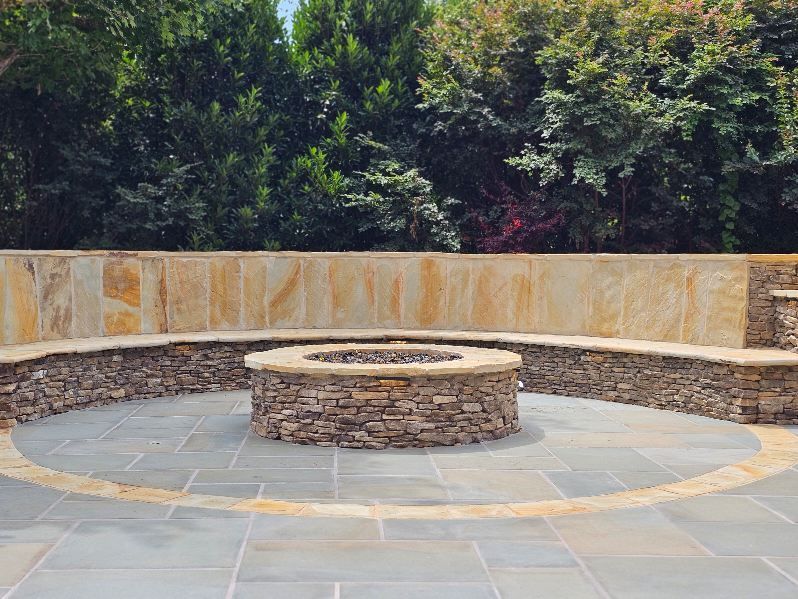 Stone fire pit with built-in seating in a patio, surrounded by a stone wall and lush greenery.