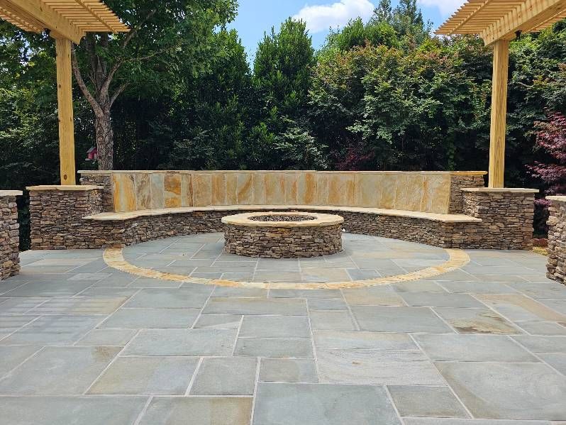 Outdoor stone patio with fire pit and built-in stone seating under a wooden pergola.
