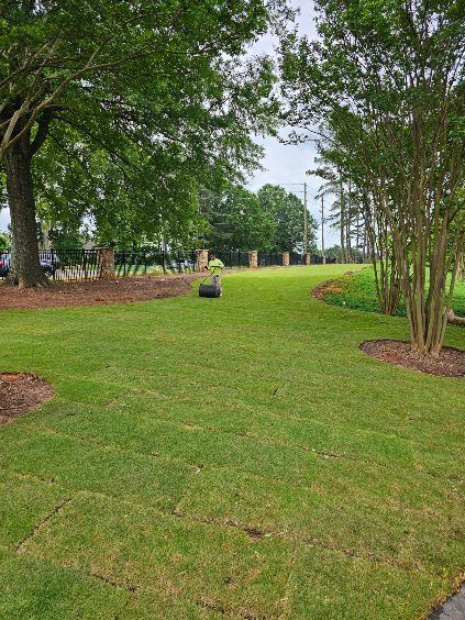 Lawn mower cutting grass in a green, open field, lined with trees and landscaping. Cloudy sky.