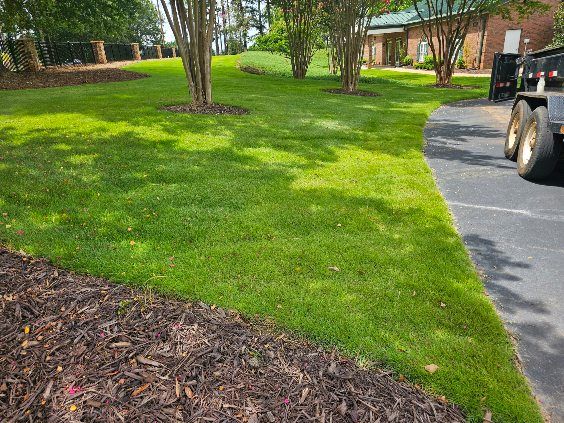 Lush green lawn with trees, mulch bed, and a paved driveway. A trailer is parked on the edge.
