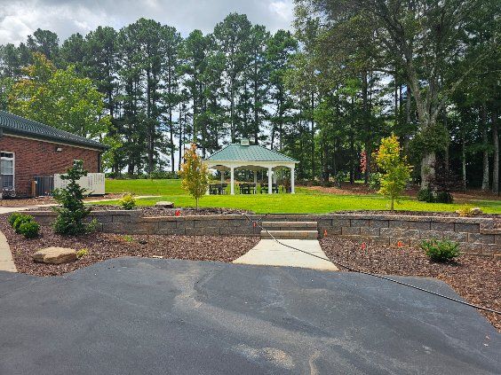 Gazebo in a grassy yard, surrounded by trees and a retaining wall. Asphalt driveway in foreground.