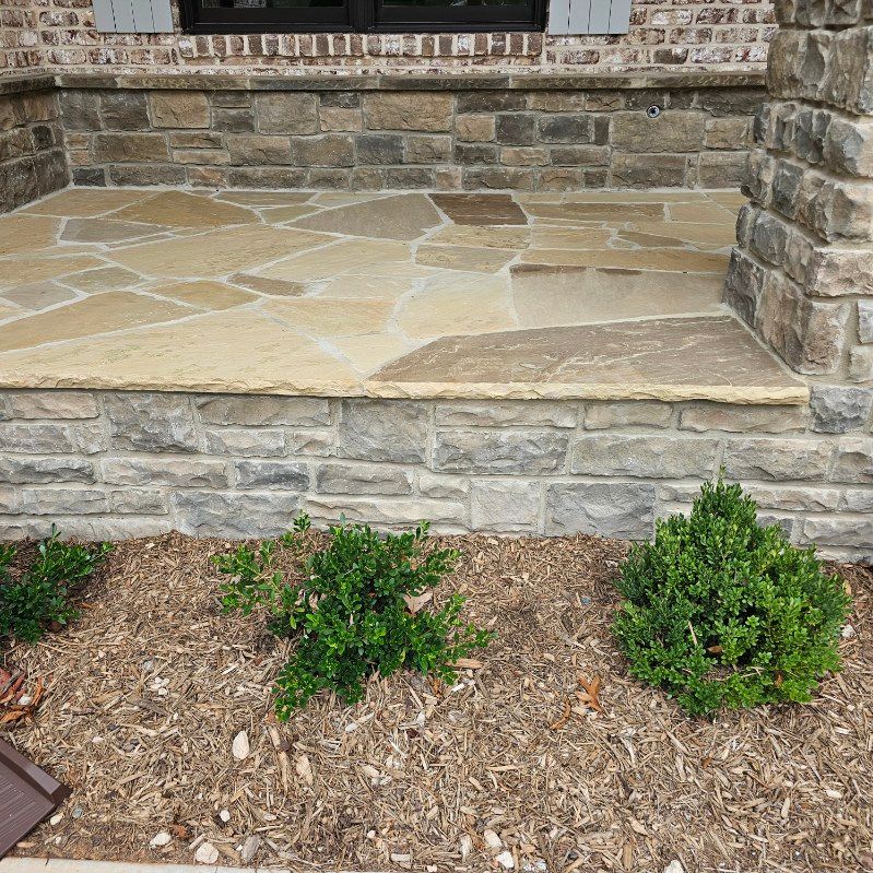 Stone porch with beige flagstone flooring, gray brick wall, and two green bushes in mulch.