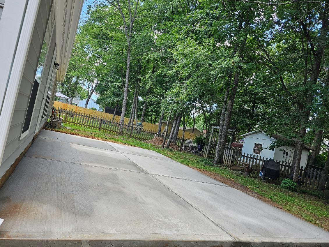 A concrete driveway leads to a fenced backyard with trees and a small white building.