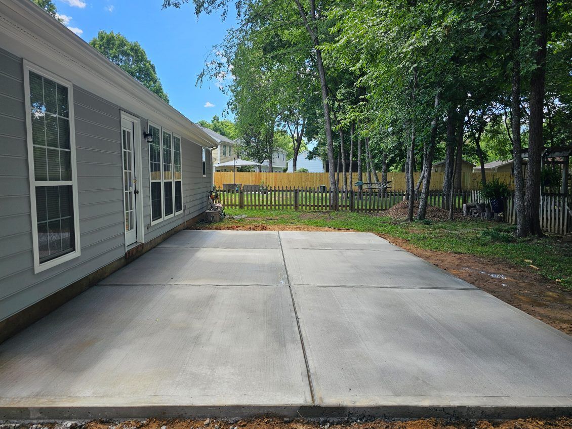 Gray concrete patio next to a light gray house, with a wooded backyard.
