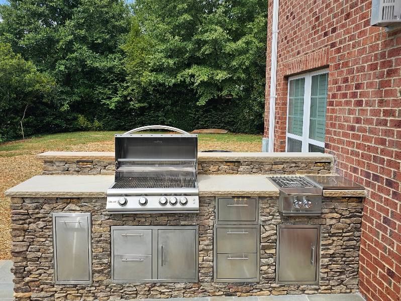 Outdoor kitchen with stainless steel grill, cabinets, and stone facade next to a brick wall.
